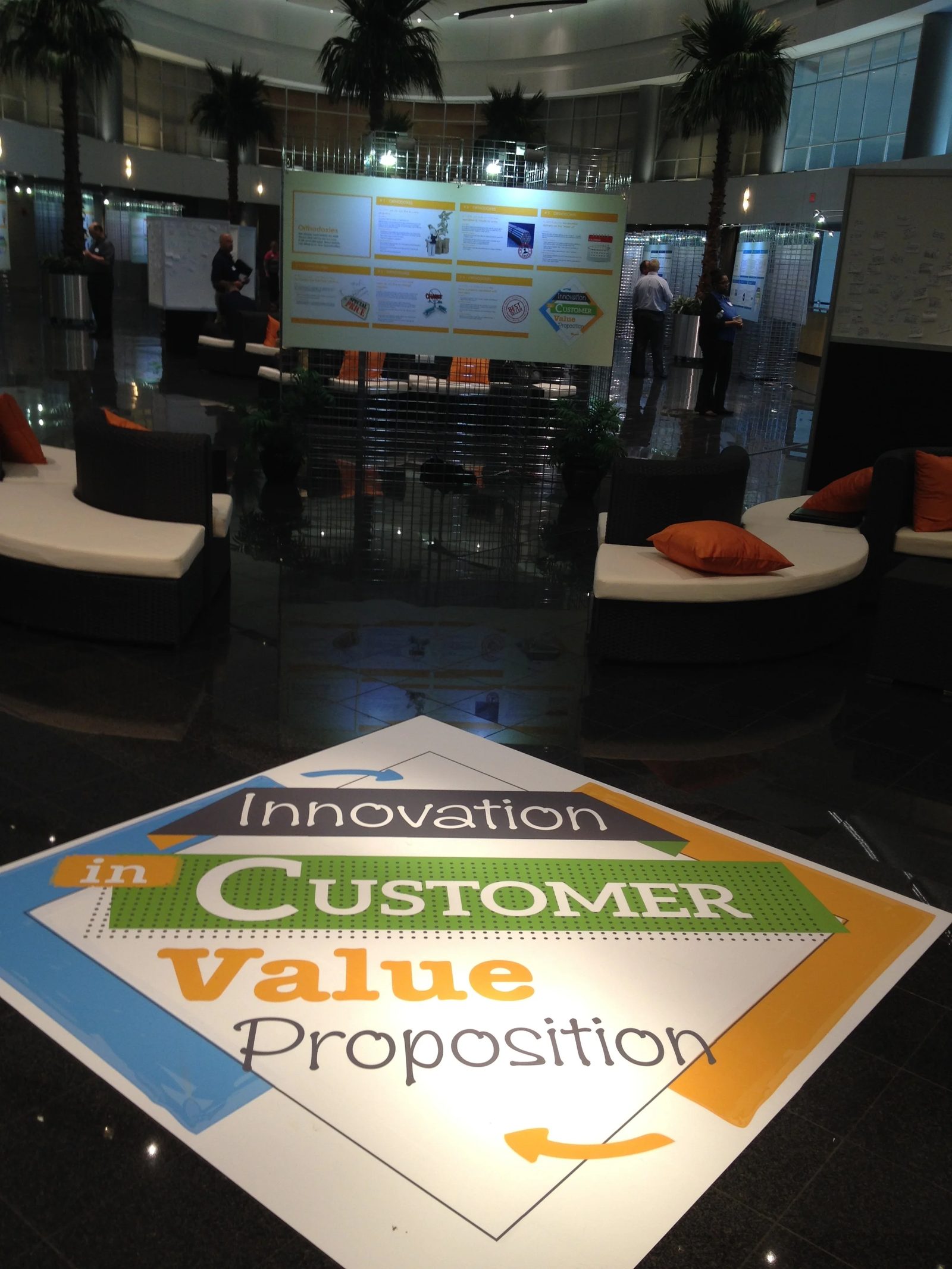 Corporate lobby at an 'Innovation in Customer Value Proposition' fair, with a large floor-level signage diamond and project posters displayed in the background.