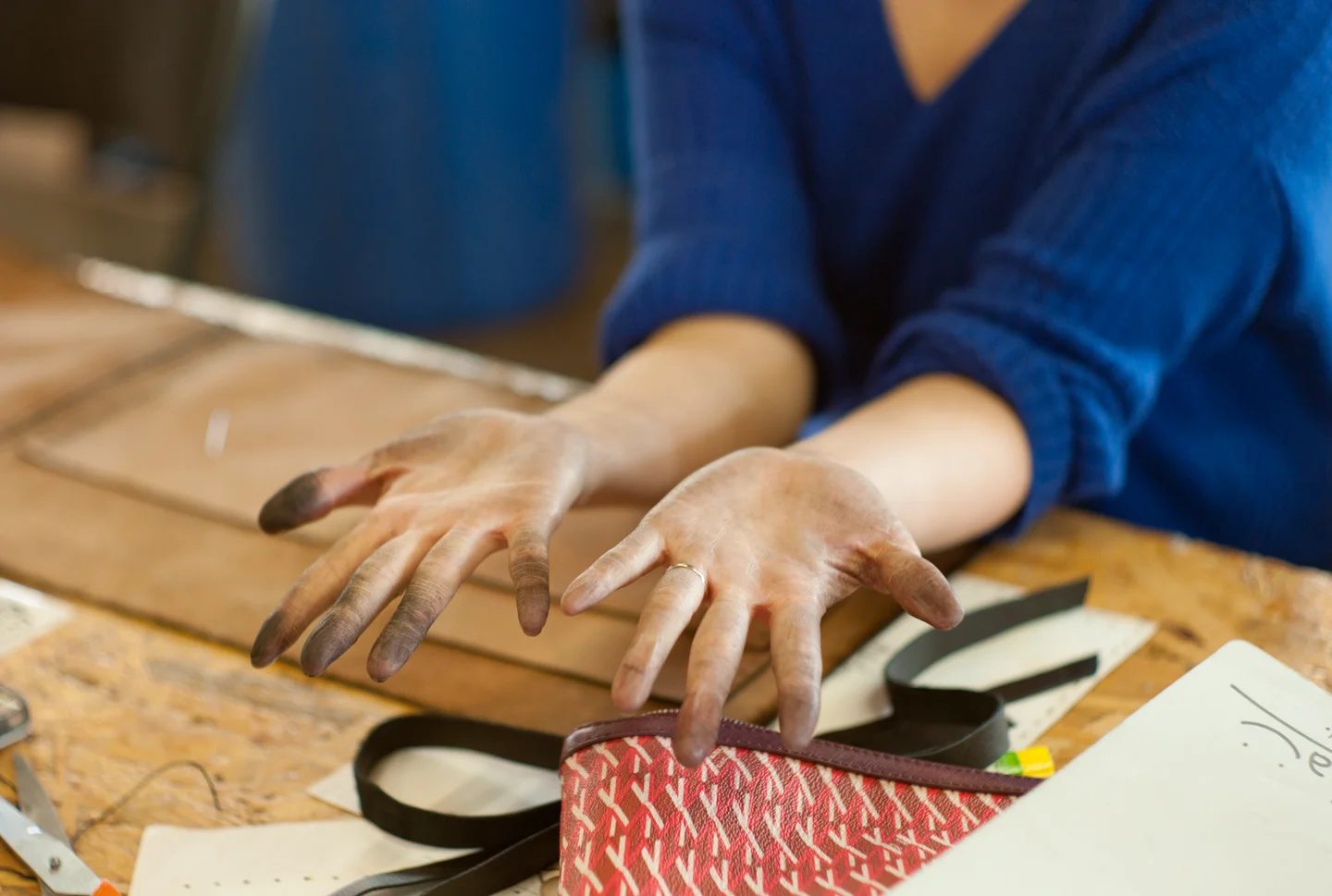 Ink-stained hands resting on a workbench scattered with leather pieces, straps, and a hand-made zippered pouch, the marks of hands-on making.